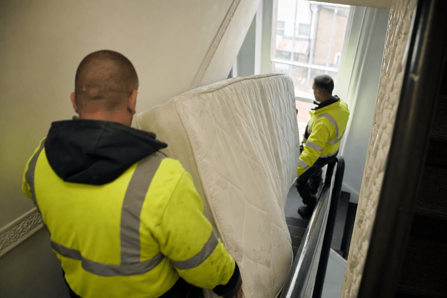 Two waste removal workers carrying a double mattress down stairs in a London flat